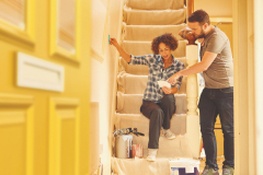 A young couple doing up their hallway in the old house they have bought . They are sitting on the dust sheets on the stairs and looking through the paint sample cards choosing the final colour for the hall . The man is holding them to the wall as the woman decides .